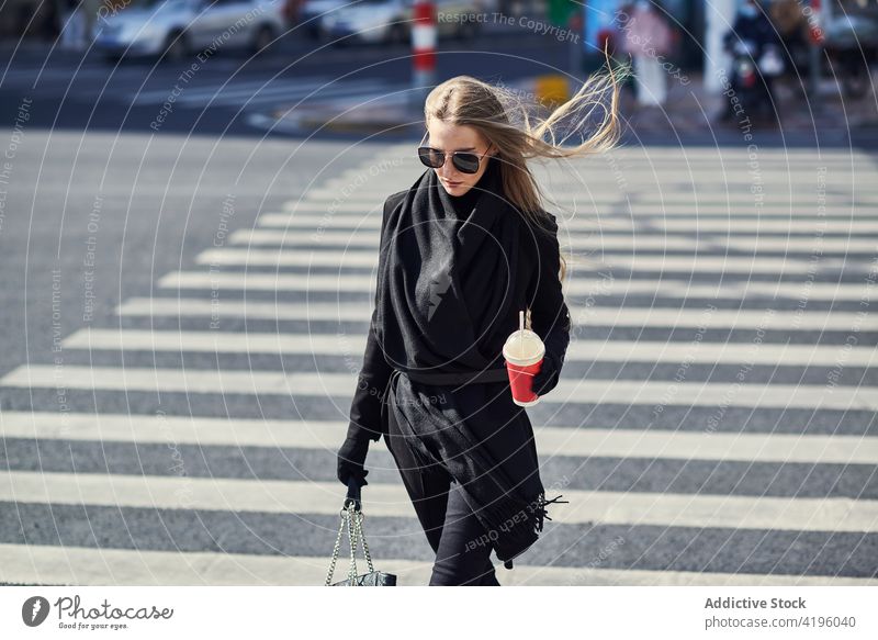 Stilvolle Frau mit erfrischendem Getränk zum Mitnehmen beim Überqueren einer städtischen Straße stylisch fliegendes Haar trinken schlendern Mode Stadt Lifestyle