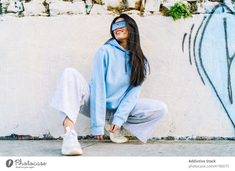 Stilvolle lächelnde Frau mit Sonnenbrille auf städtischem Bürgersteig Mode Individualität cool Zeitgenosse Lächeln Straßenbelag Großstadt Kniebeuge Kapuzenpulli