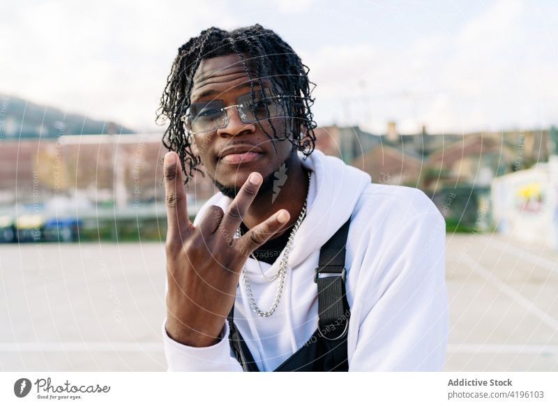 Cooler ethnischer Mann zeigt Hip-Hop-Geste auf der Straße Rap cool stylisch Hipster Individualität modern Porträt gestikulieren zeigen Vollbart Afro-Look