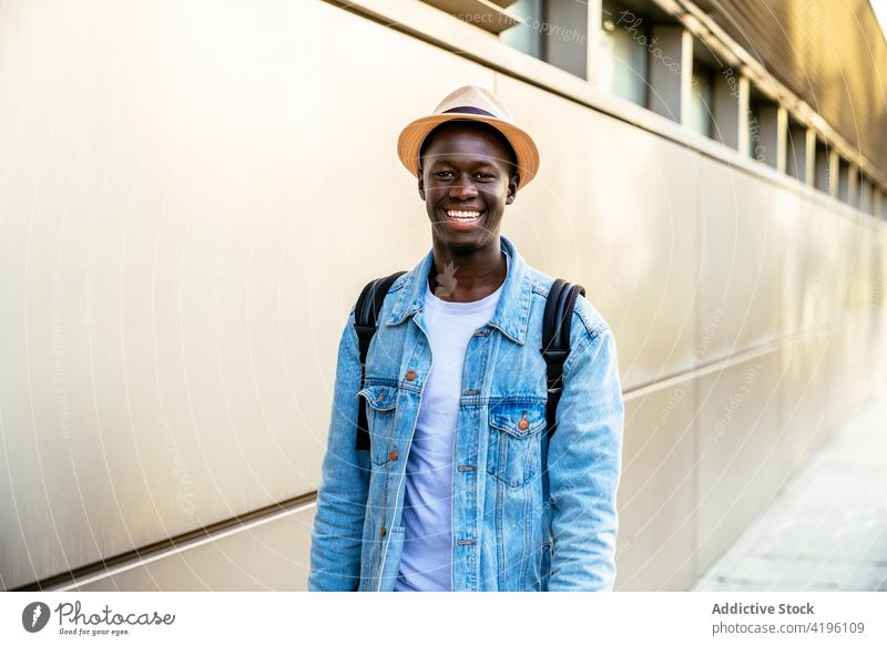 Afroamerikanischer Mann lächelnd an der Wand heiter herzlich lässig Stil freundlich Glück genießen Hand an der Hüfte Jeansstoff Jacke maskulin hell modern Stoff