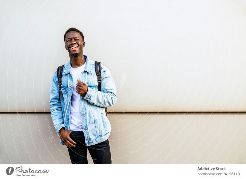 Lächelnder afroamerikanischer Mann mit Hand an der Hüfte in der Nähe der Wand heiter herzlich lässig Stil freundlich Glück genießen Jeansstoff Jacke maskulin