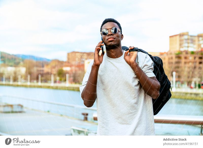 Schwarzer Mann mit modischer Sonnenbrille, der am Ufer mit seinem Smartphone spricht reden maskulin Macho trendy Stauanlage Großstadt benutzend Apparatur