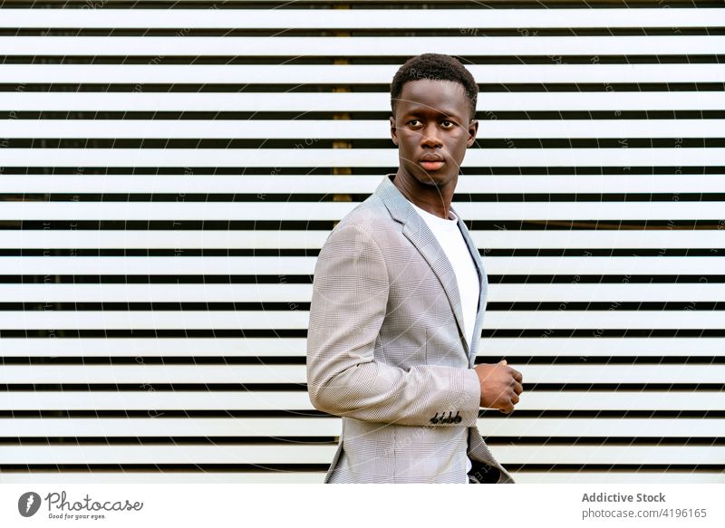 Stilvoller schwarzer Mann auf städtischem Bürgersteig in der Nähe einer gerippten Wand stylisch Mode maskulin Macho achtsam Model auf Zehenspitzen Stadt trendy