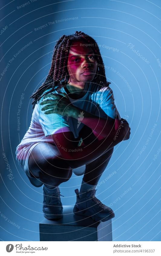 Ernstes schwarzes Teenager-Mädchen sitzt auf den Hüften in einem Neon-Studio selbstbewusst Stil Körperhaltung Youngster cool neonfarbig Persönlichkeit