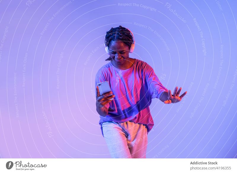 Schwarze Teenager-Tänzerin mit Smartphone im Kopfhörer im Studio Mädchen Licht Körperhaltung leuchten Youngster neonfarbig cool glühen Generation urban Atelier