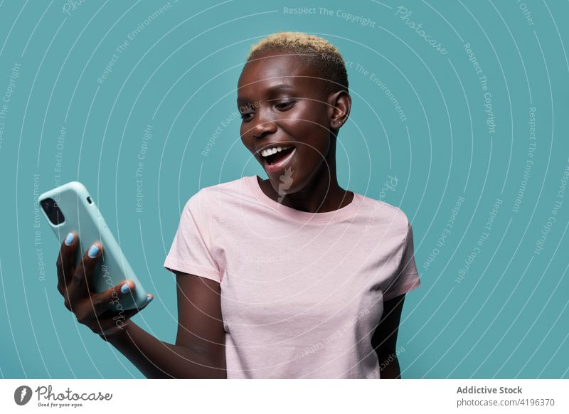 Fröhliche schwarze Frau, die im Studio ein Mobiltelefon benutzt Smartphone expressiv Afrikanisch trendy feminin Studioaufnahme Zahnfarbenes Lächeln Handy Gerät