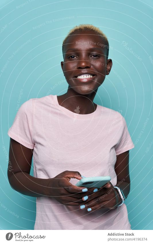 Fröhliche schwarze Frau, die im Studio ein Mobiltelefon benutzt Smartphone expressiv Afrikanisch trendy feminin Studioaufnahme Zahnfarbenes Lächeln Handy Gerät