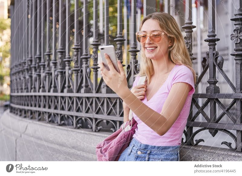 Lächelnde blonde Teenagerin beim Surfen auf dem Smartphone an einen Zaun gelehnt Frau benutzend positiv sorgenfrei Browsen digital Freizeit Internet Gerät