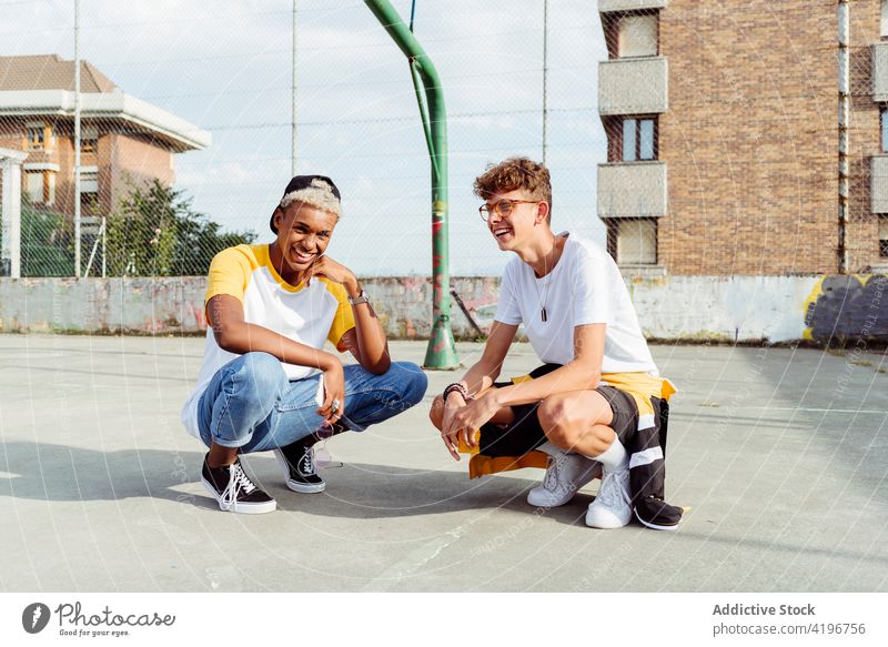 Zwei Teenager hocken auf dem städtischen Basketballplatz und lachen. Herbst schwarz Junge Kaukasier heiter Großstadt farbenfroh Gesellschaft Tageslicht ethnisch