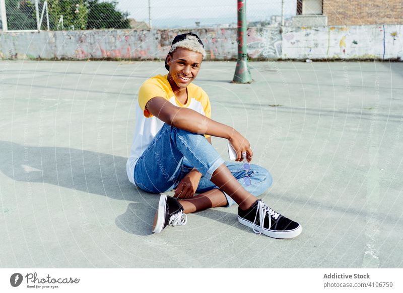 Hübscher junger schwarzer Mann sitzt und lacht auf dem Boden auf der Straße attraktiv Junge Verschlussdeckel heiter Großstadt farbenfroh Tageslicht ethnisch