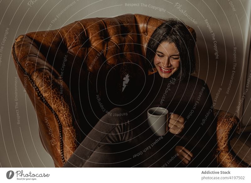 Fröhliche Frau mit Tasse im Sessel trinken gemütlich Armsessel Lächeln heimwärts genießen Komfort Getränk Freude heiß sitzen froh Wochenende jung Glee Kaffee