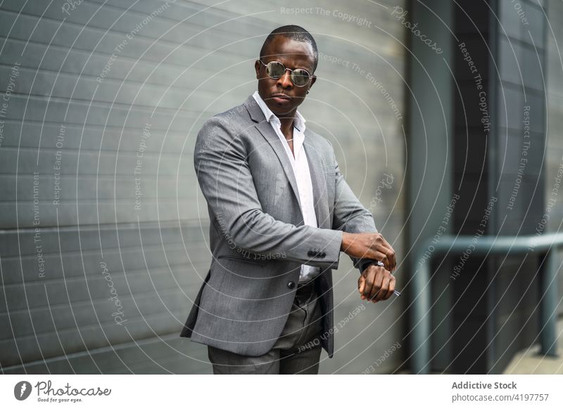 Stilvoller ethnischer Unternehmer, der die Zeit vor einer grauen Wand beobachtet zuschauend Afroamerikaner schwarz Mann schwarzpünktlich Mode gut gekleidet