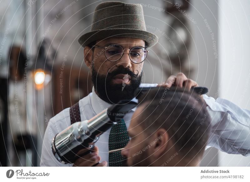Ethnischer Friseur trocknet das Haar eines Kunden im Friseursalon Haartrockner Klient Schönheit Verfahren maskulin Hipster viril elektrisch Barbershop Männer
