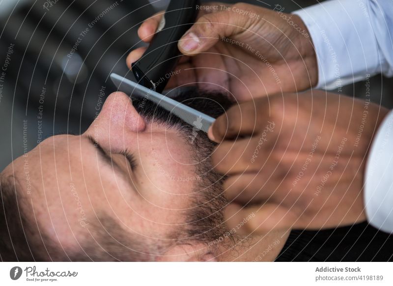 Ethnischer Barbier beim Trimmen des Schnurrbarts eines Kunden in einem Friseursalon Klient geschnitten Trimmer Kamm Pflege Verfahren Schönheit Barbershop Männer