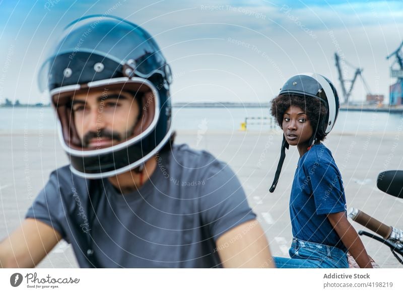 Positives junges gemischtrassiges Paar mit Helmen sitzt in der Nähe eines Motorrads am Meer Biker Seeküste Spazierweg Stil Zusammensein Termin & Datum