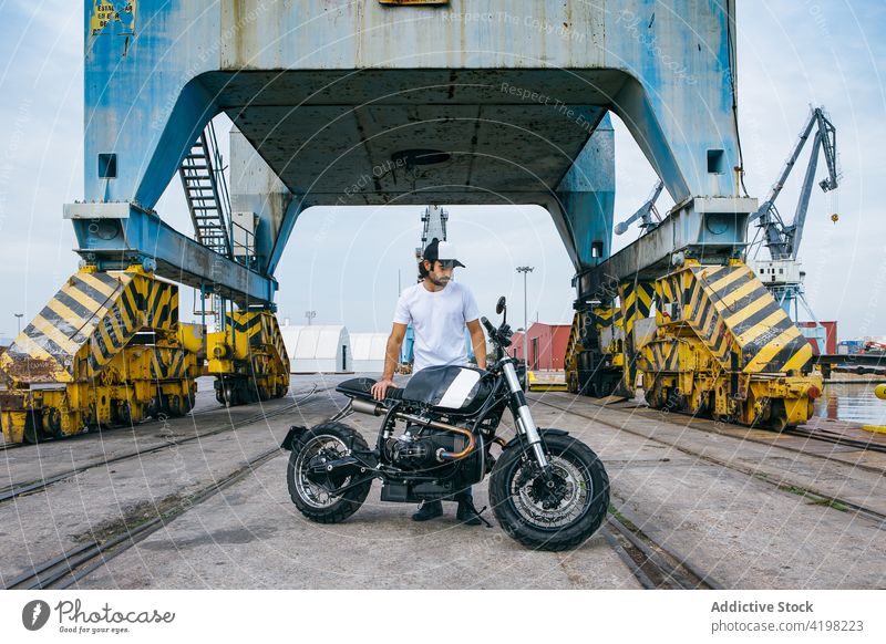 Cal junger ethnischer Mann mit Motorrad in der Nähe von Kränen, die in der Nähe des Hafens arbeiten Biker Portwein Kranich industriell ernst brutal Verkehr