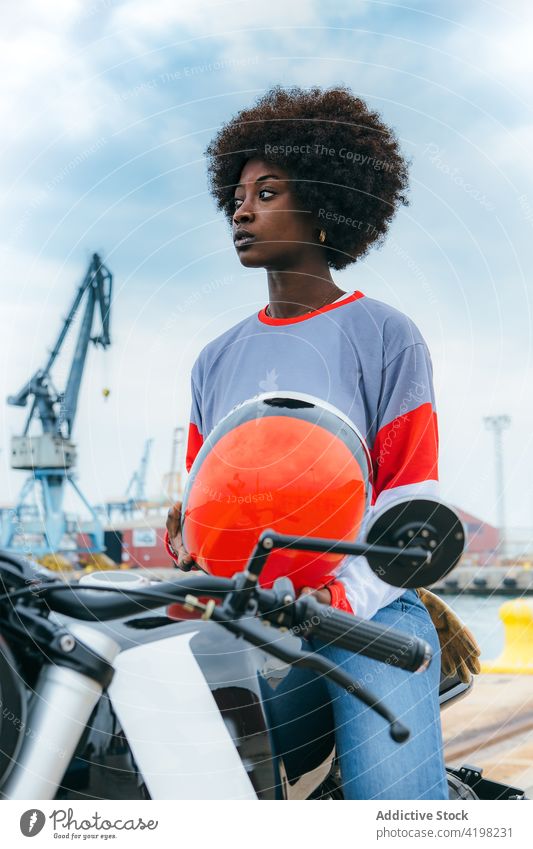 Seriöse junge ethnische Frau mit Helm auf dem Motorrad sitzend Seeküste Vorschein Persönlichkeit trendy Stauanlage allein Biker cool reisen Fahrzeug schwarz