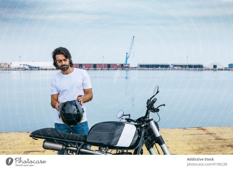Ernster junger ethnischer Mann auf einem Motorrad an einer Böschung Schutzhelm Seeküste selbstbewusst brutal ernst Stauanlage Stil Biker lässig männlich T-Shirt
