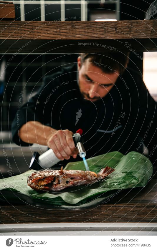 Koch bereitet Meeresfrüchte auf Palmblatt zu Mann Küchenchef Gasfackel Fisch Brandwunde Blatt Handfläche kulinarisch heiß männlich Lebensmittel Mahlzeit lecker