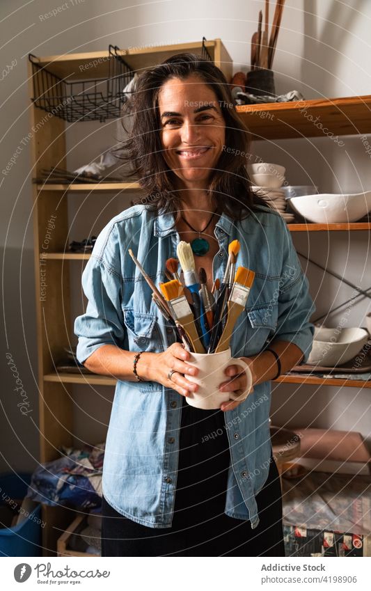 Glückliche Handwerkerin hält Tasse mit verschiedenen Pinseln in der Werkstatt Frau Lächeln Pinselblume Meister Töpferwaren Tonwaren heiter Kunstgewerbler