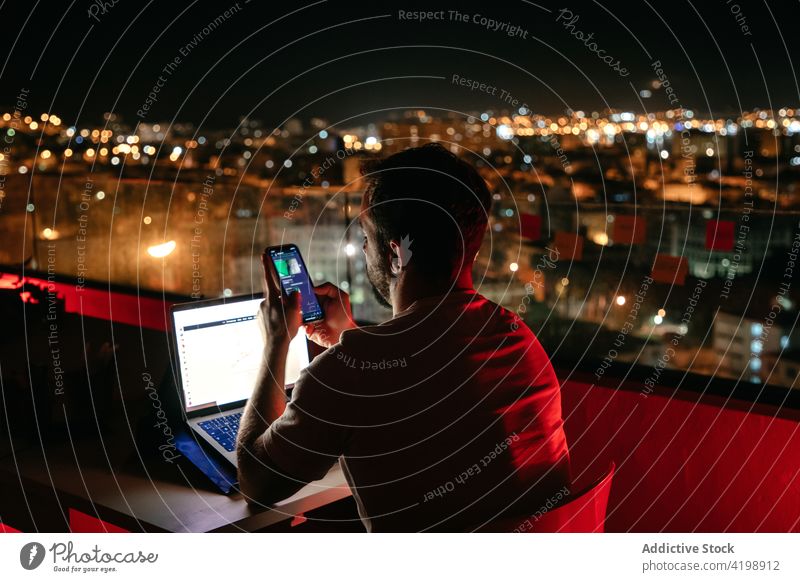 Konzentrierter Mann mit Smartphone und Laptop bei nächtlicher Online-Arbeit auf dem Dach Nachricht abgelegen Dachterrasse Abend freiberuflich Konzentration