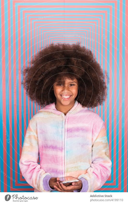 Erfreutes ethnisches Mädchen mit Smartphone im Studio stehend Afro-Look Frisur Atelier Lächeln heiter Teenager farbenfroh lebhaft schwarz Afroamerikaner Wand