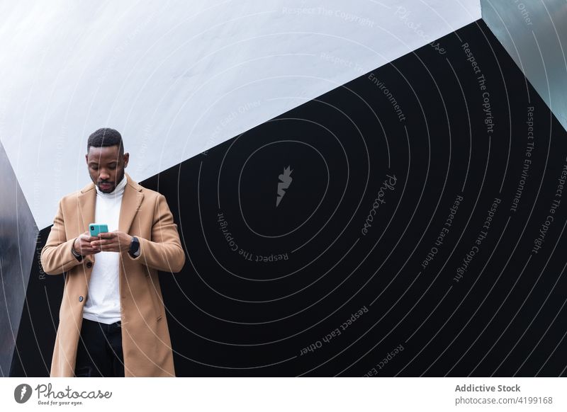 Gut gekleideter schwarzer Mann, der in der Nähe der Wand mit seinem Smartphone spricht Talkrunde Internet online achtsam maskulin Macho benutzend Apparatur