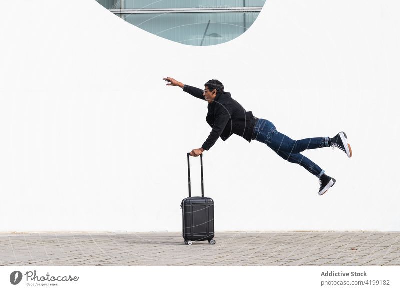 Ethnischer Tourist mit Koffer springt in die Luft über städtischem Pflaster Reisender springen sich[Akk] beeilen Eile Fliege Arm angehoben stylisch Straßenbelag