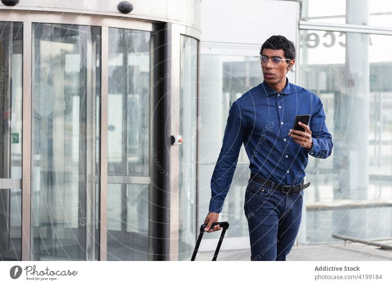 Stilvoller ethnischer Geschäftsmann mit Smartphone und Gepäck vor einer Glaswand smart lässig maskulin achtsam reisen Ausflug Lifestyle Mann Jeanshose Gurt Hemd