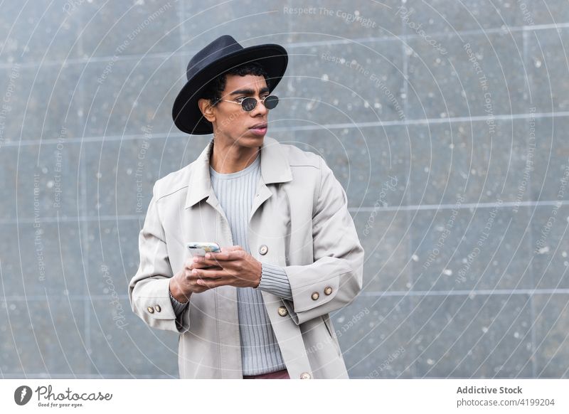Stilvoller ethnischer Mann mit Smartphone auf der Straße genießen trendy Drahtlos benutzend Apparatur Gerät Internet online Funktelefon Hut Kleidungsstück Mode
