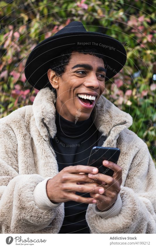 Stilvoller glücklicher ethnischer Mann mit Smartphone auf der Straße genießen trendy Drahtlos benutzend Apparatur Gerät Internet online Funktelefon Hut