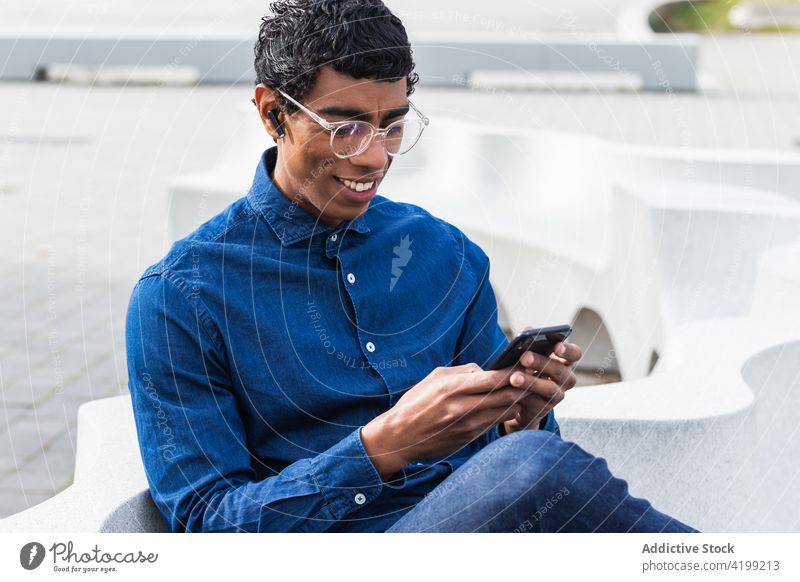 Aufmerksamer ethnischer Geschäftsmann, der in der Stadt mit seinem Smartphone chattet Unternehmer Textnachrichten Lächeln Internet Glück online interessiert