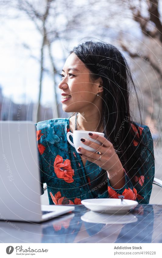 Langhaarige brünette asiatische Frau, die auf der Terrasse eines Cafés einen Kaffee trinkt, während sie mit einem Laptop arbeitet Menschen Werkstatt Chinesisch