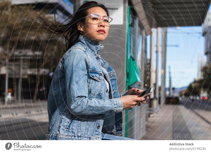 Langhaarige brünette asiatische Frau, die ein Mobiltelefon benutzt, während sie auf einer Bank auf der Straße sitzt Funktelefon Chinesisch Mobile Telefon