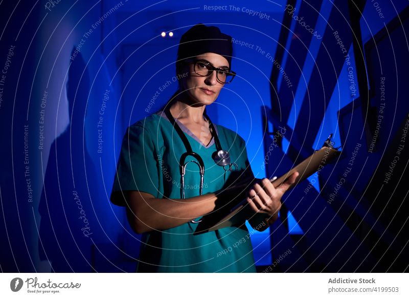 Seriöser Sanitäter mit Klemmbrett im dunklen Krankenhaus Frau Arzt Zwischenablage dunkel Klinik neonfarbig blau Licht Uniform Stethoskop leuchten Treppenhaus