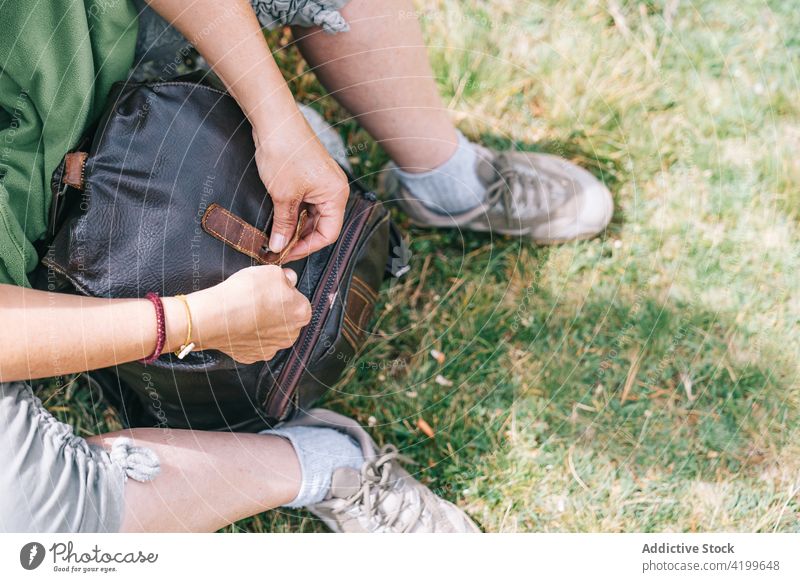 Crop gesichtslose Frau schließt Rucksack auf grasbewachsener Lichtung schließen offen Waldwiese Backpacker Grasland Trekking Camping Pause ruhen sitzen sonnig