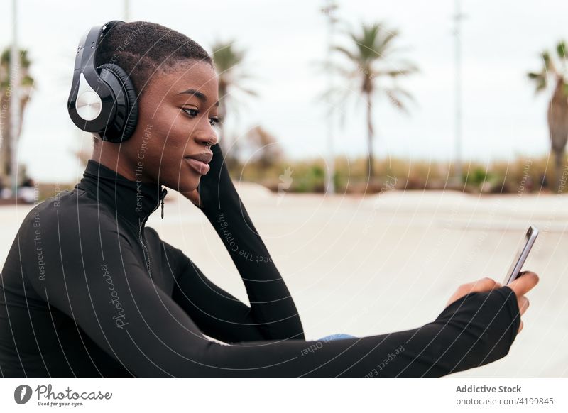 Lächelnde schwarze Frau mit Headset beobachtet Smartphone auf der Straße zuschauend Afroamerikaner Gesang zuhören Kurze Haare Inhalt Internet Porträt benutzend
