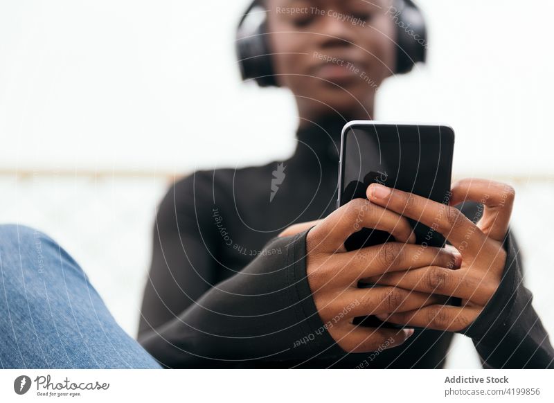 Lächelnde schwarze Frau mit Headset beobachtet Smartphone auf der Straße zuschauend Afroamerikaner Gesang zuhören Kurze Haare Inhalt Internet Porträt benutzend