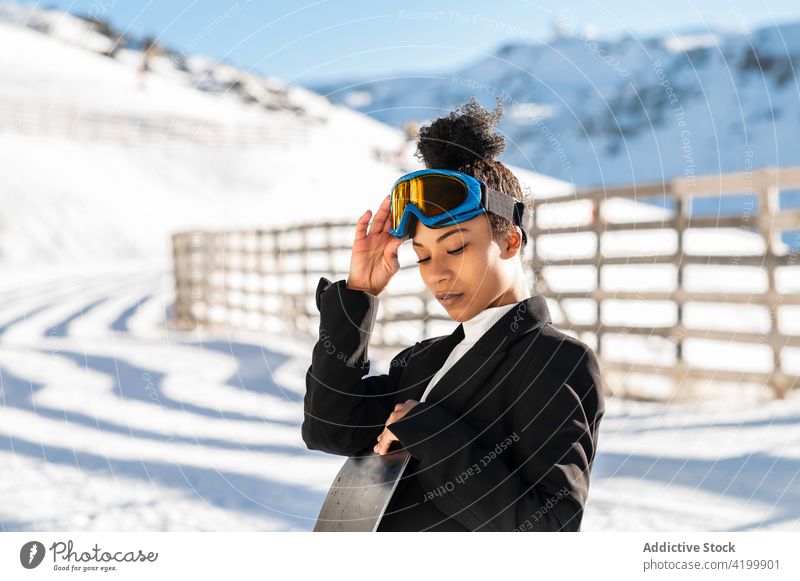 Stilvolle schwarze Sportlerin mit Schneeball auf Snowboard Winter stylisch Brille freundlich Frau genießen zeigen Anzug trendy Stiefel Winterzeit kalt Wetter