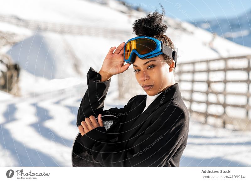 Stilvolle schwarze Sportlerin mit Schneeball auf Snowboard Winter stylisch Brille freundlich Frau genießen zeigen Anzug trendy Stiefel Winterzeit kalt Wetter