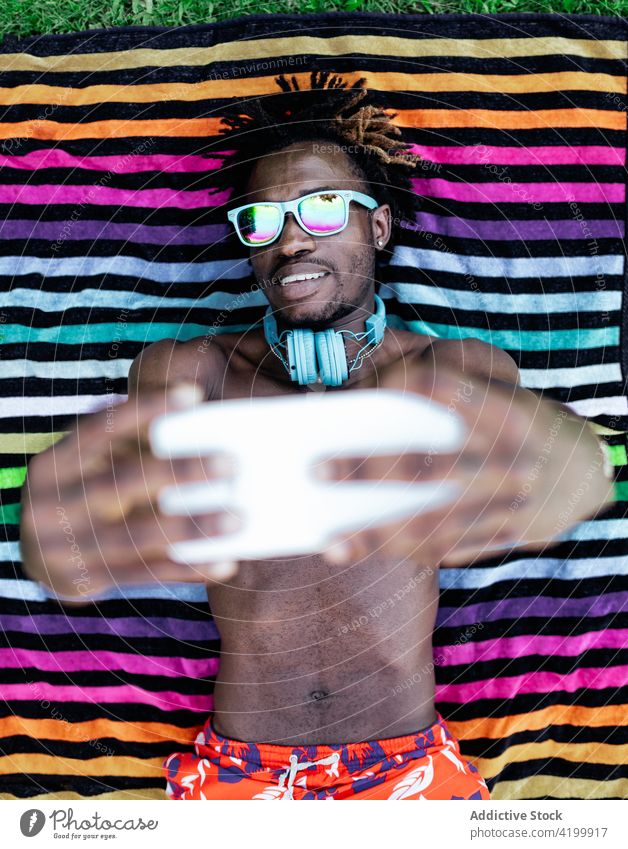 Ethnischer Mann, der auf einem Handtuch liegt und ein Selfie macht Smartphone Sommer Gras Lügen Selbstportrait genießen Urlaub männlich ethnisch schwarz