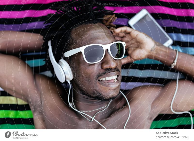 Fröhlicher schwarzer Mann, der mit Kopfhörern Musik hört zuhören Handtuch Sommer Rasen heiter Gesang genießen männlich Afroamerikaner ethnisch sorgenfrei Lügen