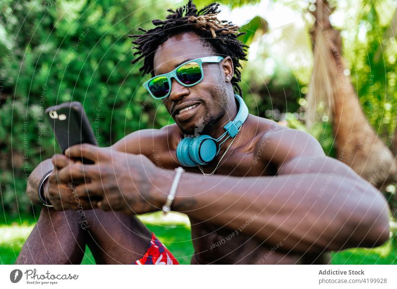 Schwarzer Mann benutzt Smartphone im Sommerpark Browsen Urlaub nackter Torso Nachricht Sonnenbrille Wiese Park männlich ethnisch schwarz Afroamerikaner