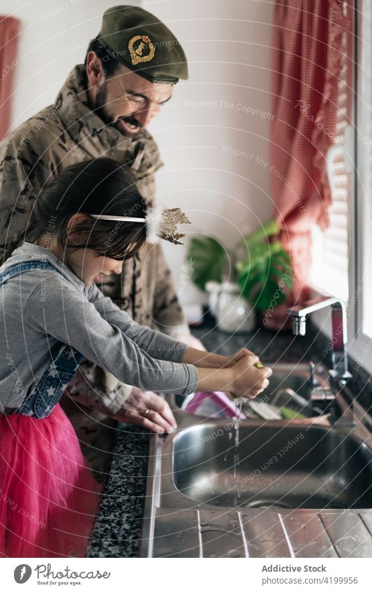 Lächelnder Militärvater und süßes Mädchen waschen gemeinsam Geschirr Vater Tochter Soldat Zusammensein Küche Waschen heimwärts Waschbecken Abfertigungsschalter