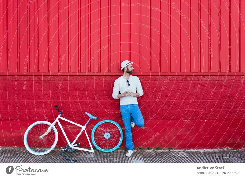 Mann mit Smartphone steht in der Nähe von Fahrrad gegen rotes Gebäude sich[Akk] entspannen Straße trendy Stil rote Wand ruhen Mode nachdenklich Großstadt sonnig