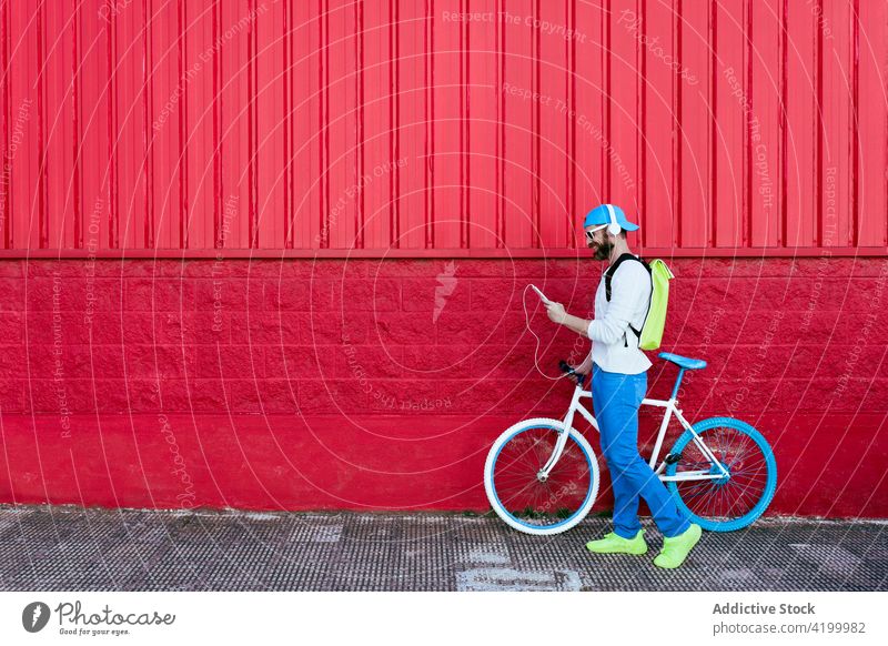 Mann mit Smartphone und Kopfhörern geht mit Fahrrad an rotem Gebäude vorbei Straße trendy Stil rote Wand ruhen Mode Großstadt urban männlich Vollbart cool