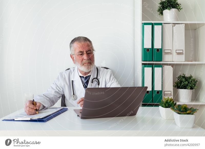 Älterer Arzt macht sich während eines Videogesprächs auf dem Laptop in der Klinik Notizen arzt Videoanruf schreiben Gesundheitswesen Beratung Internet abgelegen