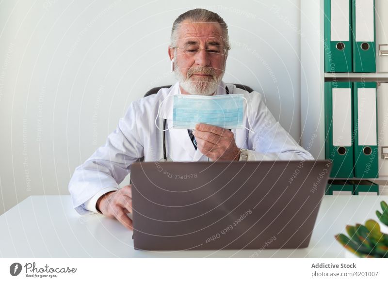 Oberarzt zeigt während eines Videogesprächs eine sterile Maske gegen den Laptop Arzt Mundschutz Videoanruf Gesundheitswesen Beratung online behüten Mann
