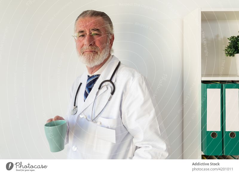 Älterer Arzt mit einer Tasse Kaffee in der Klinik Uniform professionell Spezialist Stethoskop Mann Porträt organisieren Heißgetränk Getränk Tee Brille