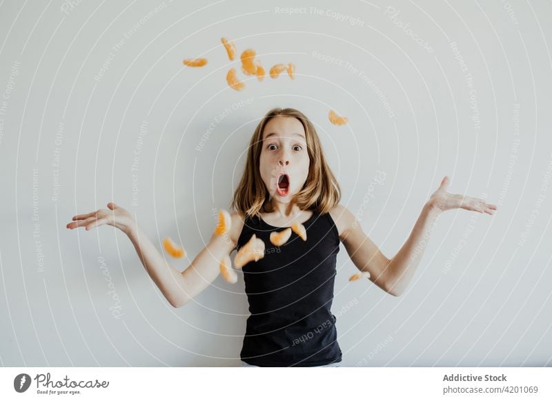 Aufgeregtes Mädchen, das Mandarinenscheiben wirft und eine Grimasse gegen eine weiße Wand zieht Kind werfen Mund geöffnet erstaunt Schock Spaß haben